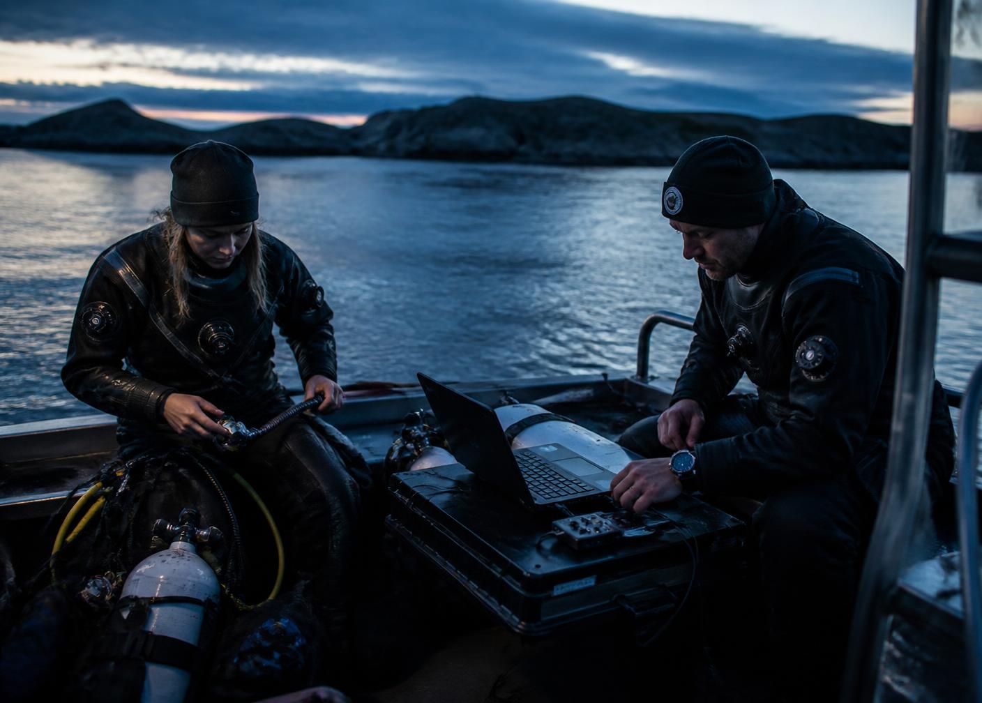 Two divers preparing gear and logging data on a boat at dusk